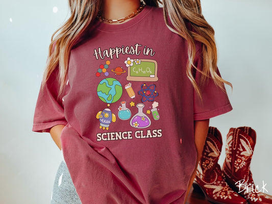 A woman wearing a cute, vintage brick colored Comfort Colors T-shirt with the text Happiest in Science Class. In between the text are a globe, chemistry, rocket ship, chalkboard, science beakers and test tubes, a pencil, DNA molecules, and Saturn.