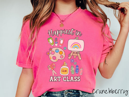 A woman wearing a cute, vintage crunchberry colored Comfort Colors T-shirt with the text Happiest in Art Class. In between the text are paint brushes, a paint palette, an art canvas on a stand, scissors, glue, crayons, and flowers.