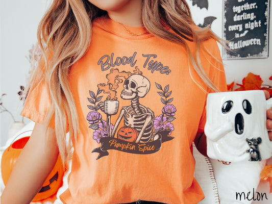 A woman wearing a cute, vintage melon colored Comfort Colors T-shirt with text Blood Type: Pumpkin Spice. There is a skeleton holding a pumpkin spiced latte with orange steam coming out and a pumpkin while surrounded by purple flowers.