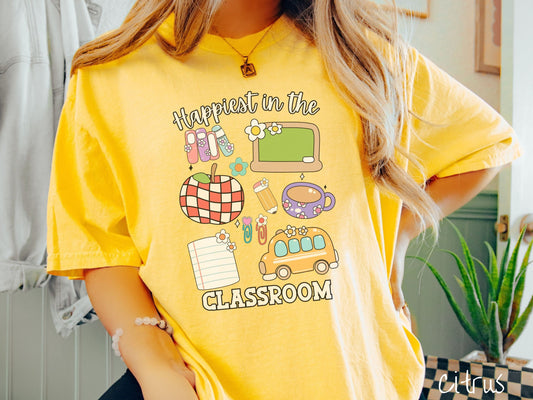 A woman wearing a cute, vintage citrus colored Comfort Colors T-shirt with the text Happiest in the Classroom. In between the text are books, an apple, chalkboard, pencil and paper, coffee cup, yellow school bus, paperclips, and flowers.