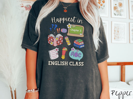 A woman wearing a cute, vintage pepper colored Comfort Colors T-shirt with the text Happiest in English Class. In between the text are books, erasers, a chalkboard, pencil, pen, notebook paper, alphabet letters, and flowers.