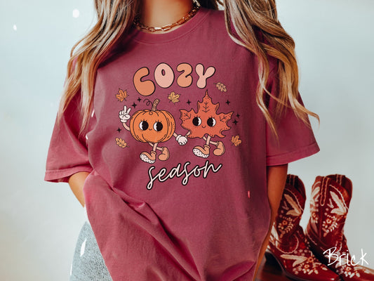 A woman wearing a cute, vintage brick colored Comfort Colors T-shirt with the text Cozy Season and in between the text are a smiling orange pumpkin holding up a peace sign with one hand and holding hands with a smiling dark orange leaf.