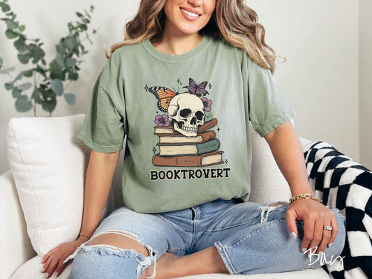 A woman wearing a cute, vintage bay colored Comfort Colors T-shirt with the text Booktrovert in black font. Above are a stack of brown and green old books with a smiling skeleton skull sitting on top with colorful butterflies and flowers.