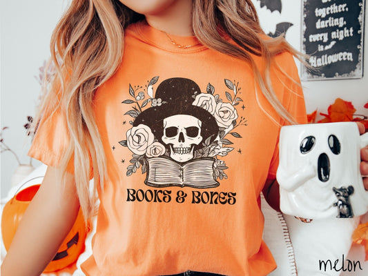 A woman wearing a cute, vintage melon colored Comfort Colors T-shirt with the text Books & Bones in black font. Above is a smiling skeleton wearing a black top hat surrounded by roses and leaves, and hovering above a thick, leather bound book.