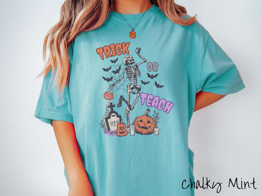 A woman wearing a cute, vintage chalky mint colored Comfort Colors T-shirt with text Trick or Teach in purple and orange font. There is a skeleton holding a pumpkin candy bucket dancing among spider webs and black bats over a tombstone and pumpkins.