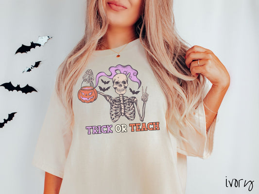 A woman wearing a cute, vintage ivory colored Comfort Colors T-shirt with text Trick or Teach in purple and orange font. Above is a skeleton holding up a peace sign and a pumpkin candy bucket with purple smoke coming out and bats in the background.