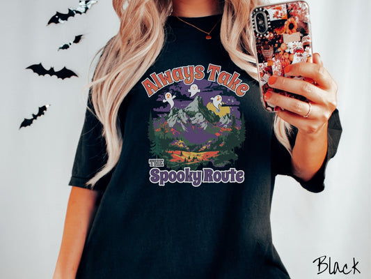 A woman wearing a cute, vintage black colored Comfort Colors T-shirt with the text Always Take the Spooky Route in colorful font. There are three white ghosts with orange faces flying above a mountain landscape with rolling hills and green forest.