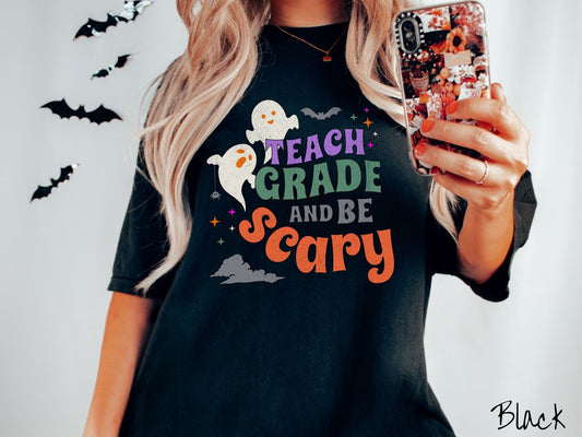 A woman wearing a cute, vintage black colored Comfort Colors T-shirt with the text Teach Grade and Be Scary in colorful font. There are orange-eyed white ghosts flying around and black bats and twinkling stars in the sky.