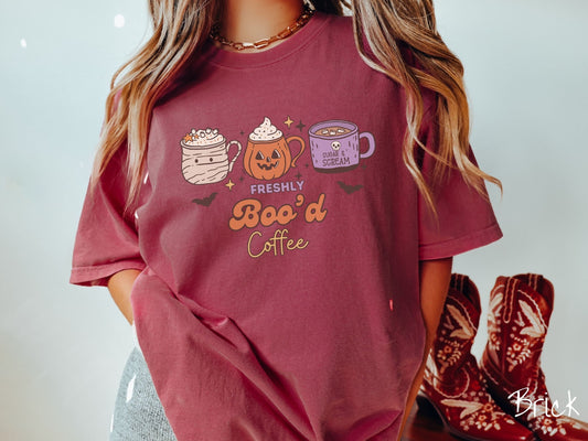 A woman wearing a cute, vintage brick colored Comfort Colors T-shirt with the text Freshly Boo’d Coffee. Above are coffee mugs where one looks like a mummy with whipped cream, one a carved pumpkin mug, and the last is purple with a skull.