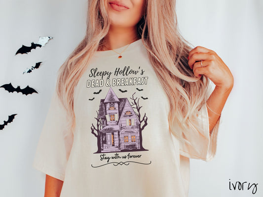 A woman wearing a cute, vintage ivory colored Comfort Colors T-shirt with the text Sleepy Hollow’s Dead & Breakfast above then Stay with us forever below. In between the text is a spooky haunted house surrounded by black bats.