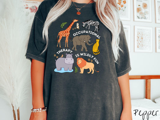 A woman wearing a cute, vintage pepper colored Comfort Colors T-shirt with the text Occupational Therapy is Wildly Fun. There is a giraffe, zebra elephant, hippopotamus, lion, leopard, and ground squirrel surrounded by leafy branches and paw prints.