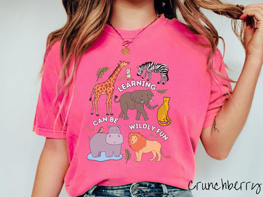 A woman wearing a cute, vintage crunchberry colored Comfort Colors T-shirt with the text Learning Can Be Wildly Fun. There is a giraffe, zebra elephant, hippopotamus, lion, leopard, and ground squirrel surrounded by leafy branches and paw prints.