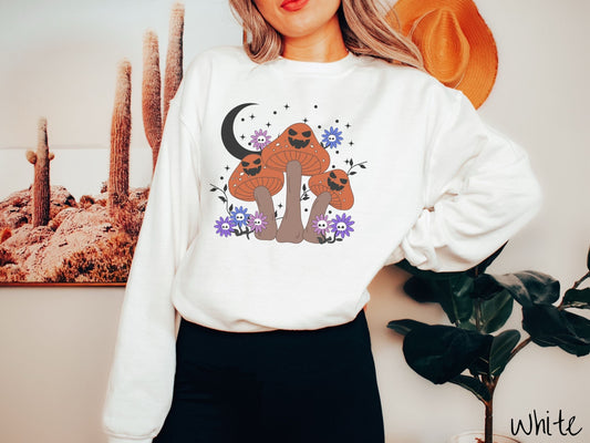 A woman wearing a cute, vintage white colored comfy Gildan sweatshirt with three dark orange and light brown mushrooms with black jack o&#39;lantern faces. They are standing around blue and purple flowers and in front of a black crescent moon.