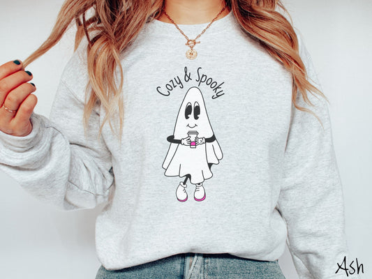 A woman wearing a cute, vintage ash colored comfy Gildan sweatshirt with the text Cozy & Spooky across the top. Below is a young, smiling white ghost holding a white and pink coffee to-go cup wearing white and pink tennis shoes.