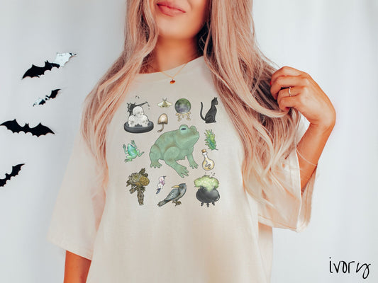 A woman wearing a cute, vintage ivory colored Comfort Colors T-shirt with a four by three grid of witches brew ingredients. There are skulls, a black cat, a giant green frog, a black crow, a bubbling cauldron, a potion flask, and some herbs.