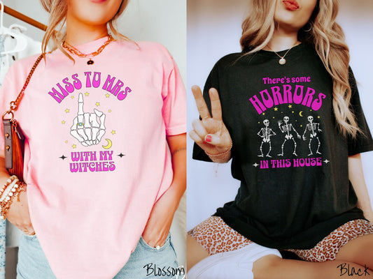 A woman wearing a vintage Comfort Colors T-shirt with the text Miss to Mrs with My Witches and a skeleton hand with a ring on it under stars. The other shirt has the text There’s some horrors in this house with skeletons dancing under the stars.