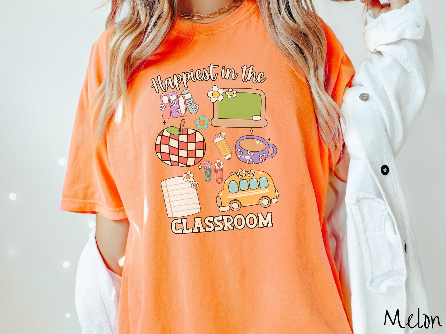 A woman wearing a cute, vintage melon colored Comfort Colors T-shirt with the text Happiest in the Classroom. In between the text are books, an apple, chalkboard, pencil and paper, coffee cup, yellow school bus, paperclips, and flowers.