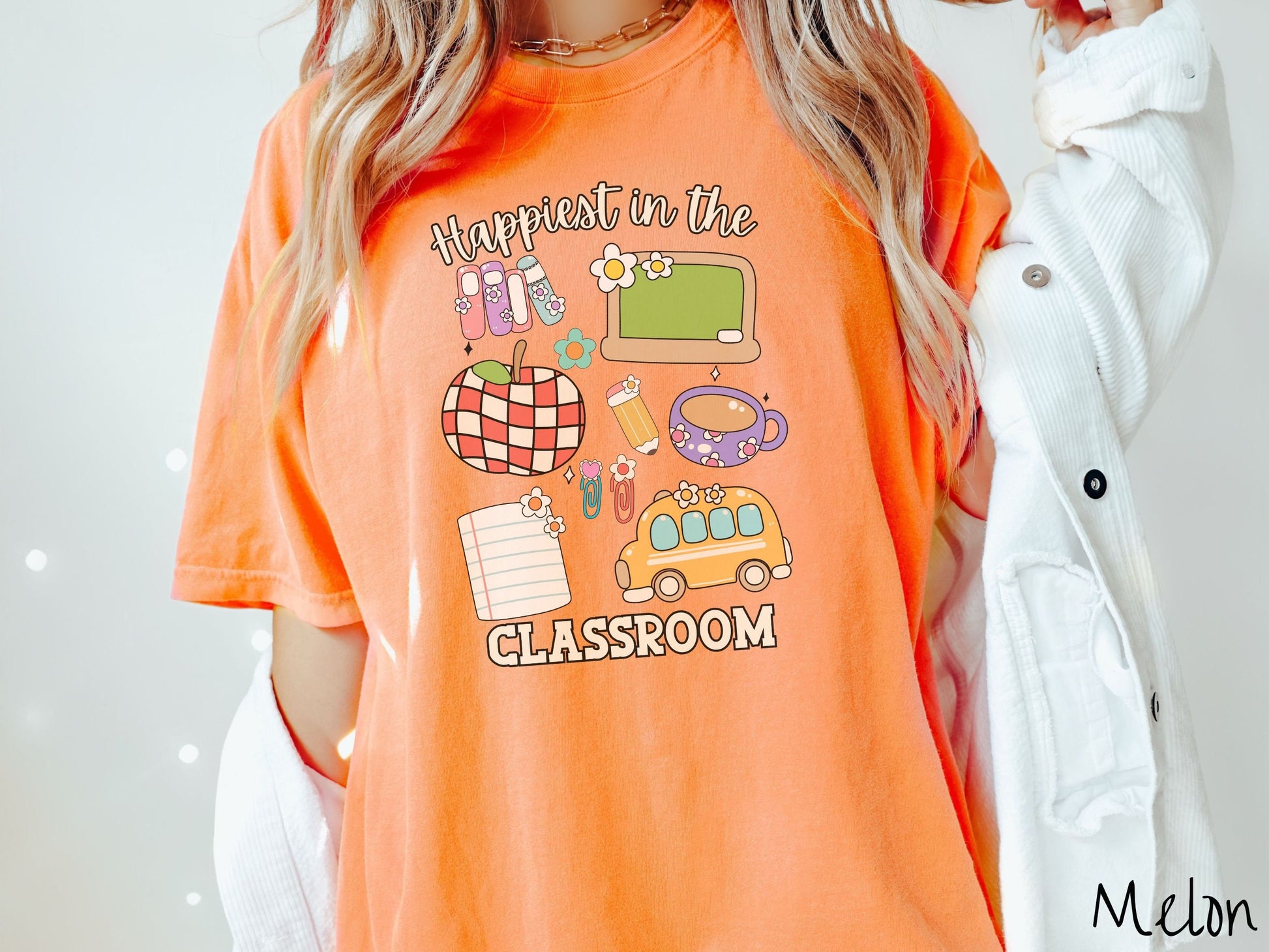 A woman wearing a cute, vintage melon colored Comfort Colors T-shirt with the text Happiest in the Classroom. In between the text are books, an apple, chalkboard, pencil and paper, coffee cup, yellow school bus, paperclips, and flowers.