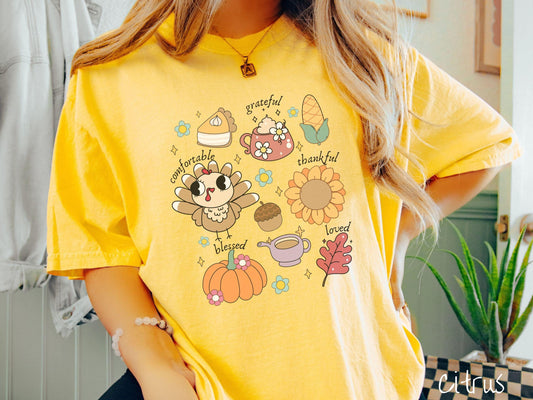 A woman wearing a cute, vintage citrus colored Comfort Colors T-shirt with text grateful comfortable thankful blessed and love. In a grid format are a piece of pumpkin pie, a coffee mug, corn, a turkey, a sunflower, a pumpkin, a leaf, and an acorn.