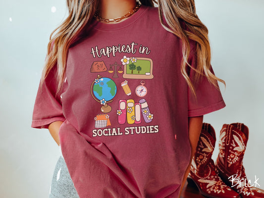 A woman wearing a cute, vintage brick colored Comfort Colors T-shirt with the text Happiest in Social Studies Class. In between the text are hieroglyphics, a globe, a pencil, compass, books, laptop computer, and a weight scale.