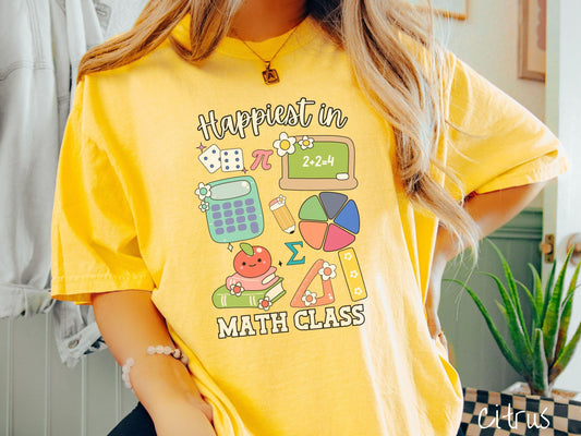 A woman wearing a cute, vintage citrus colored Comfort Colors T-shirt with the text Happiest in Math Class. In between the text are the pi symbol, a chalkboard, calculator, pencil, books, protractor, ruler, dice, an apple, and flowers.