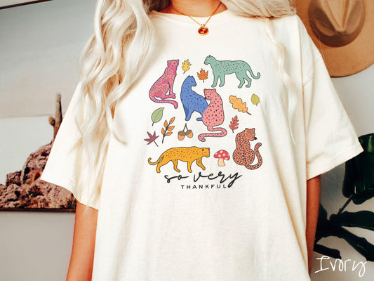 A woman wearing a cute, vintage ivory colored Comfort Colors T-shirt with the text so very thankful in black font. Above are colorful, spotted leopards in seated and walking positions, as well as leaves, acorns, and mushrooms scattered about.