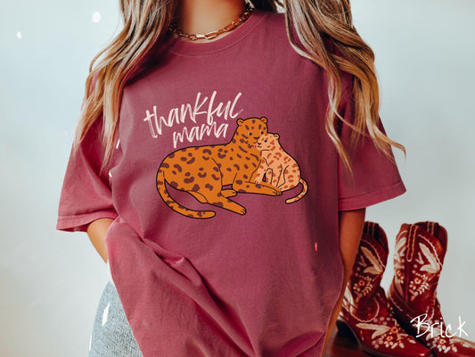 A woman wearing a cute, vintage brick colored Comfort Colors T-shirt with the text thankful mama in black, sharpie font. Below is an orange, mother leopard coddling its child.