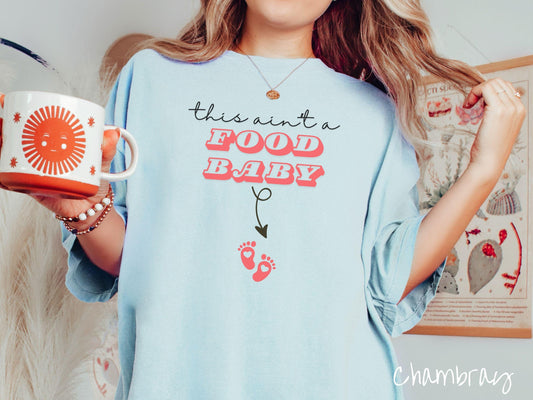 A woman wearing a cute, vintage chambray colored white colored Comfort Colors T-shirt with the text this aint a food baby in black and red font. There is a black arrow pointing down to a pair of red baby footprints with hearts in them.