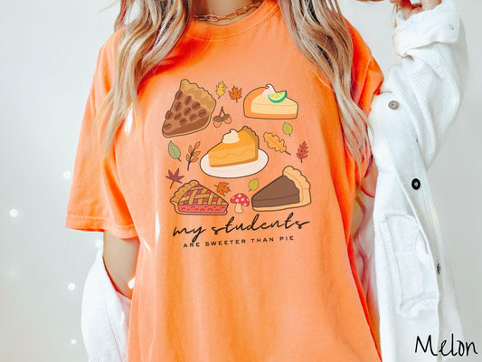 A woman wearing a cute, vintage melon colored Comfort Colors T-shirt with the text my students are sweeter than pie. Above are a piece of pecan pie, lemon meringue pie, pumpkin pie, cherry pie, and chocolate french silk pie surrounded by fall leaves.