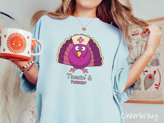 A woman wearing a cute, vintage chambray colored Comfort Colors T-shirt with the text Nursin and Turkin in white and purple font. Above is a yellow, pink, and purple turkey wearing a nursing hat with a pink cross on it.