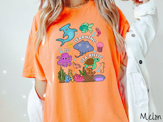 A woman wearing a cute, vintage melon colored Comfort Colors T-shirt with the text Learning is Sea-Rific. There is a shark, green fish, octopus, stingray, jellyfish, turtle, and seahorse swimming together above a colorful coral reef.