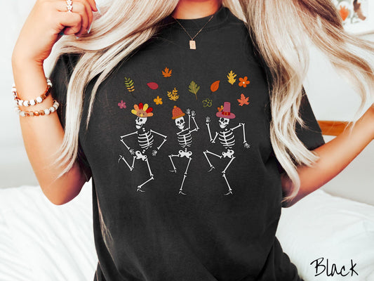 A woman wearing a cute black colored Comfort Colors t-shirt with three white skeletons wearing pilgrim hats dancing with colorful fall leaves in the air.