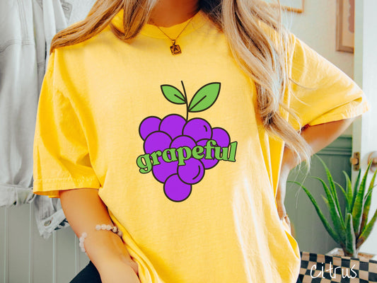 A woman wearing a cute, vintage citrus colored Comfort Colors T-shirt with the text grapeful in green font across the front. Behind grapeful is a bunch of juicy, purple grapes with a green stem.