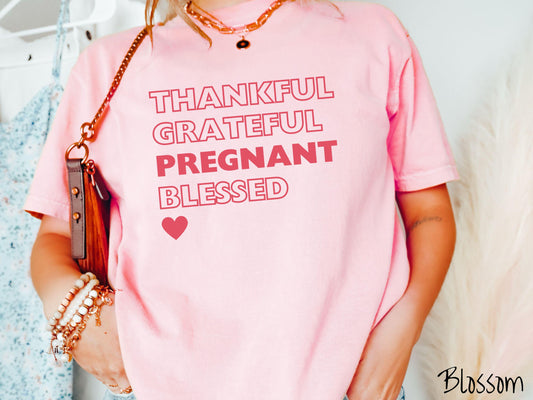 A woman wearing a cute, vintage blossom colored Comfort Colors T-shirt with the words THANKFUL GRATEFUL PREGNANT BLESSED stacked on top of each other in red font, with PREGNANT in bold font. There is also a red heart at the bottom.