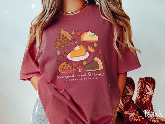 A woman wearing a cute, vintage brick colored Comfort Colors T-shirt with the text Occupational Therapy is sweeter than pie. Above are a piece of pecan pie, lemon meringue pie, pumpkin pie, cherry pie, and chocolate french silk pie.