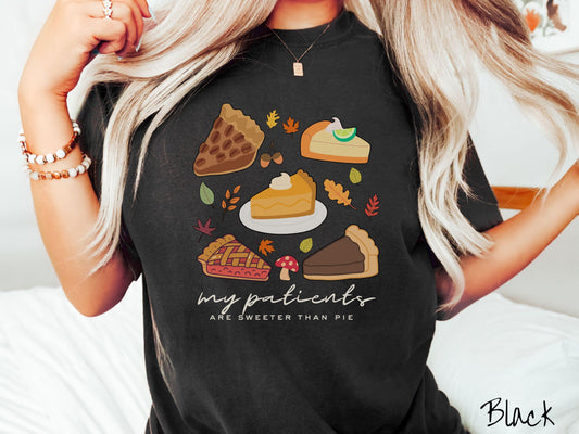 A woman wearing a cute, vintage black colored Comfort Colors T-shirt with text my patients are sweeter than pie. Above are a piece of pecan pie, lemon meringue pie, pumpkin pie, cherry pie, and chocolate french silk pie surrounded by fall leaves.