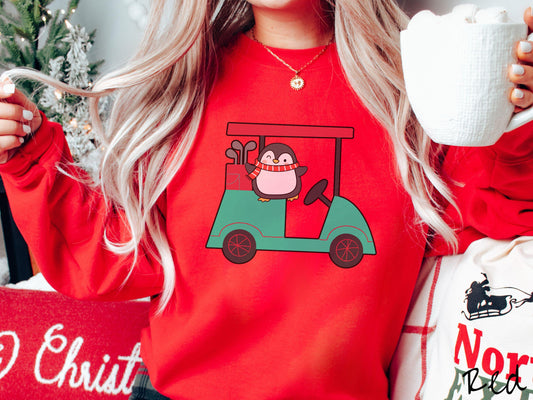 A woman wearing a cute red colored sweatshirt with smiling penguin driving a green golf cart and waving with both hands.