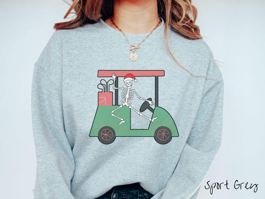 A woman wearing a cute sport grey colored sweatshirt with smiling Skeleton with a Santa Hat driving a green golf cart and waving with one hand