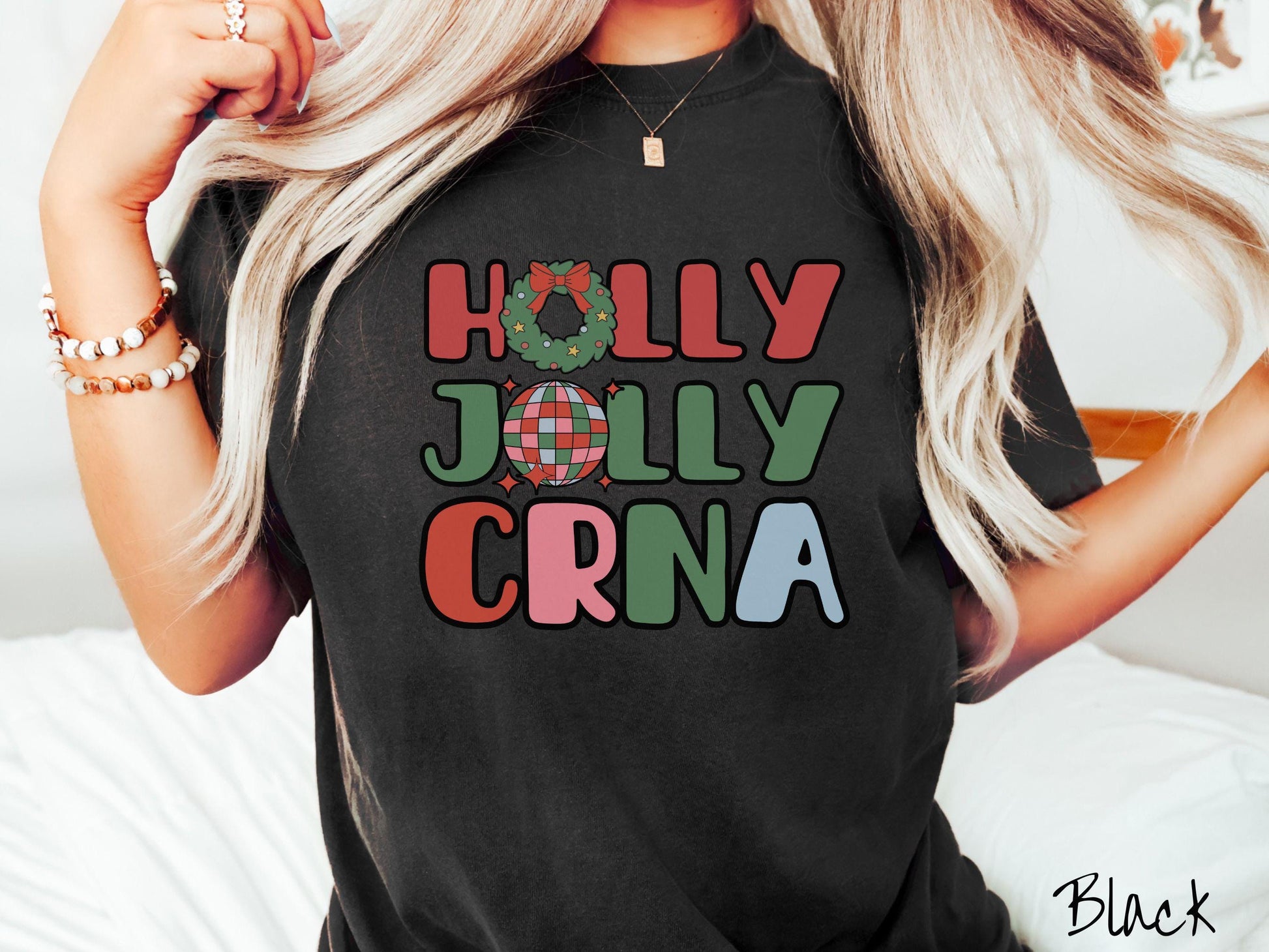 A woman wearing a cute, vintage black colored Comfort Colors T-shirt with the text Holly Jolly CRNA in red, green, pink, and blue font. The letter O in Holly is a Christmas wreath and the letter O in Jolly is a multi-colored disco ball.