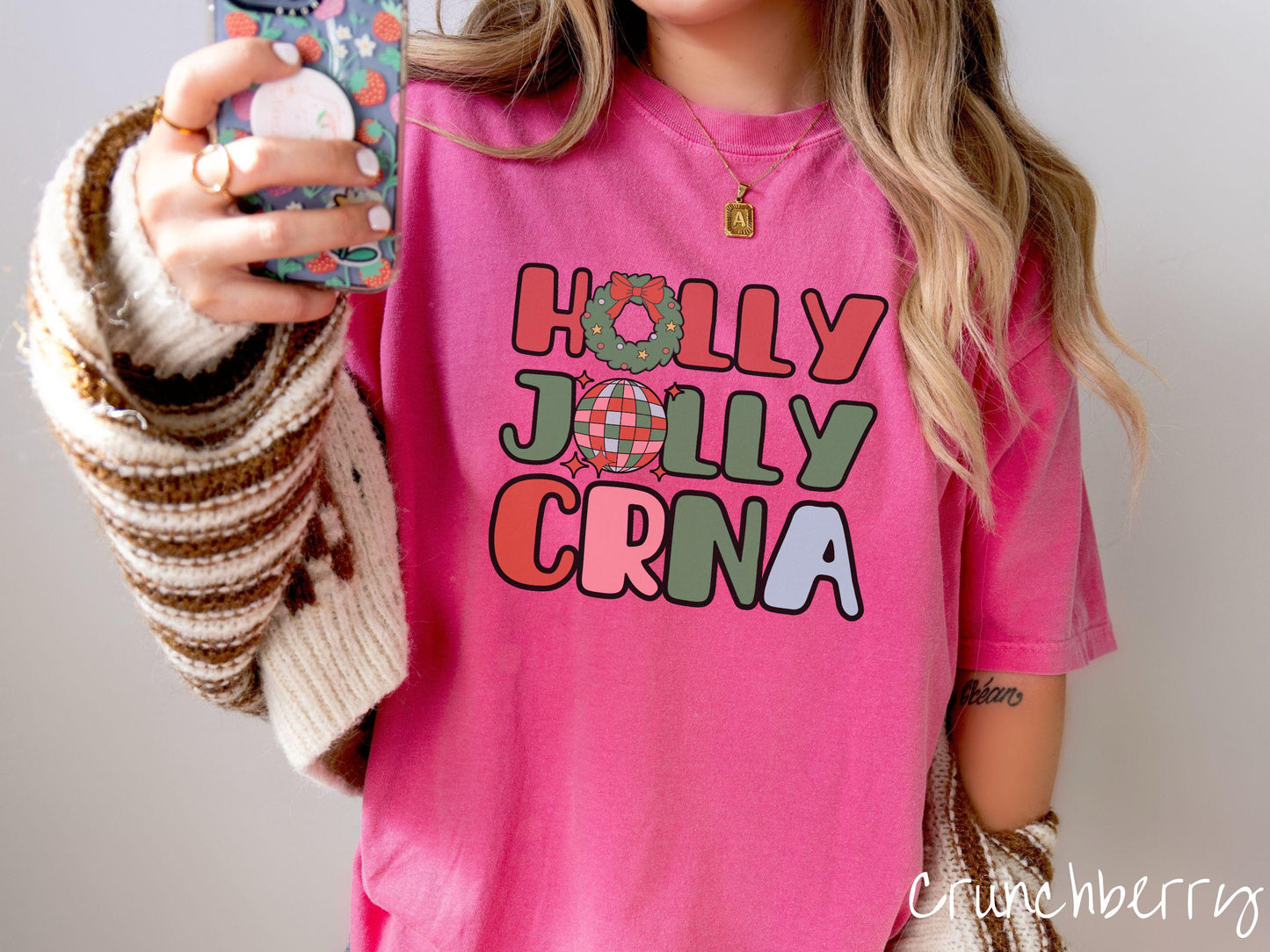A woman wearing a cute, vintage crunchberry colored Comfort Colors T-shirt with the text Holly Jolly CRNA in red, green, pink, and blue font. The letter O in Holly is a Christmas wreath and the letter O in Jolly is a multi-colored disco ball.