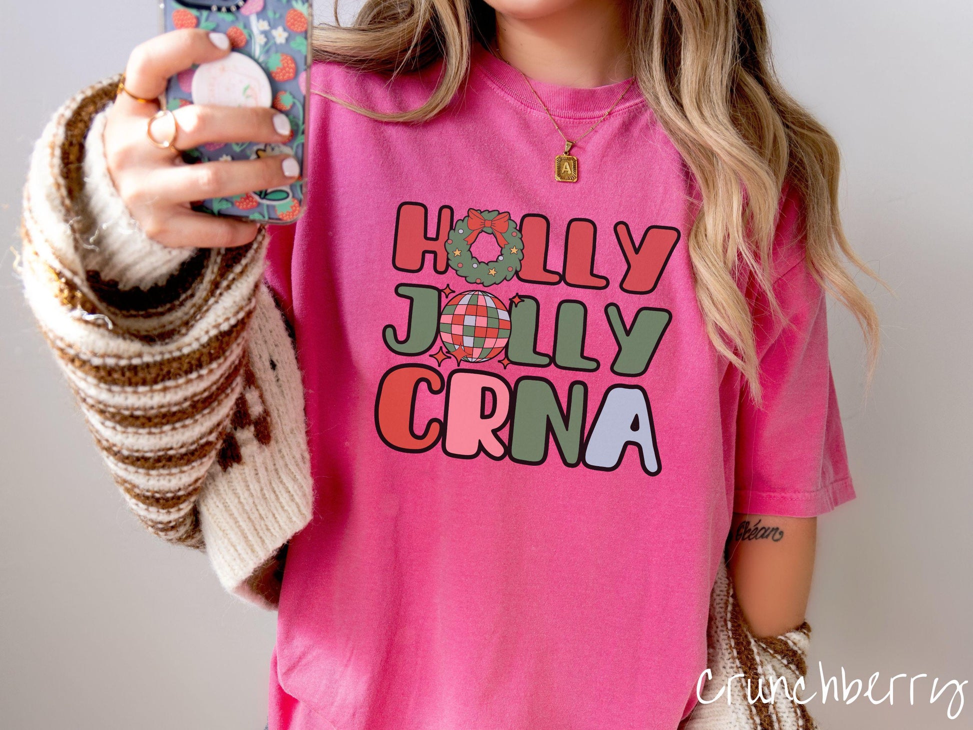 A woman wearing a cute, vintage crunchberry colored Comfort Colors T-shirt with the text Holly Jolly CRNA in red, green, pink, and blue font. The letter O in Holly is a Christmas wreath and the letter O in Jolly is a multi-colored disco ball.