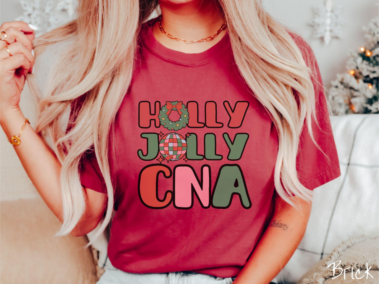 A woman wearing a cute, vintage brick colored Comfort Colors T-shirt with the text Holly Jolly CNA in red, green, pink, and blue font. The letter O in Holly is a Christmas wreath and the letter O in Jolly is a multi-colored disco ball.