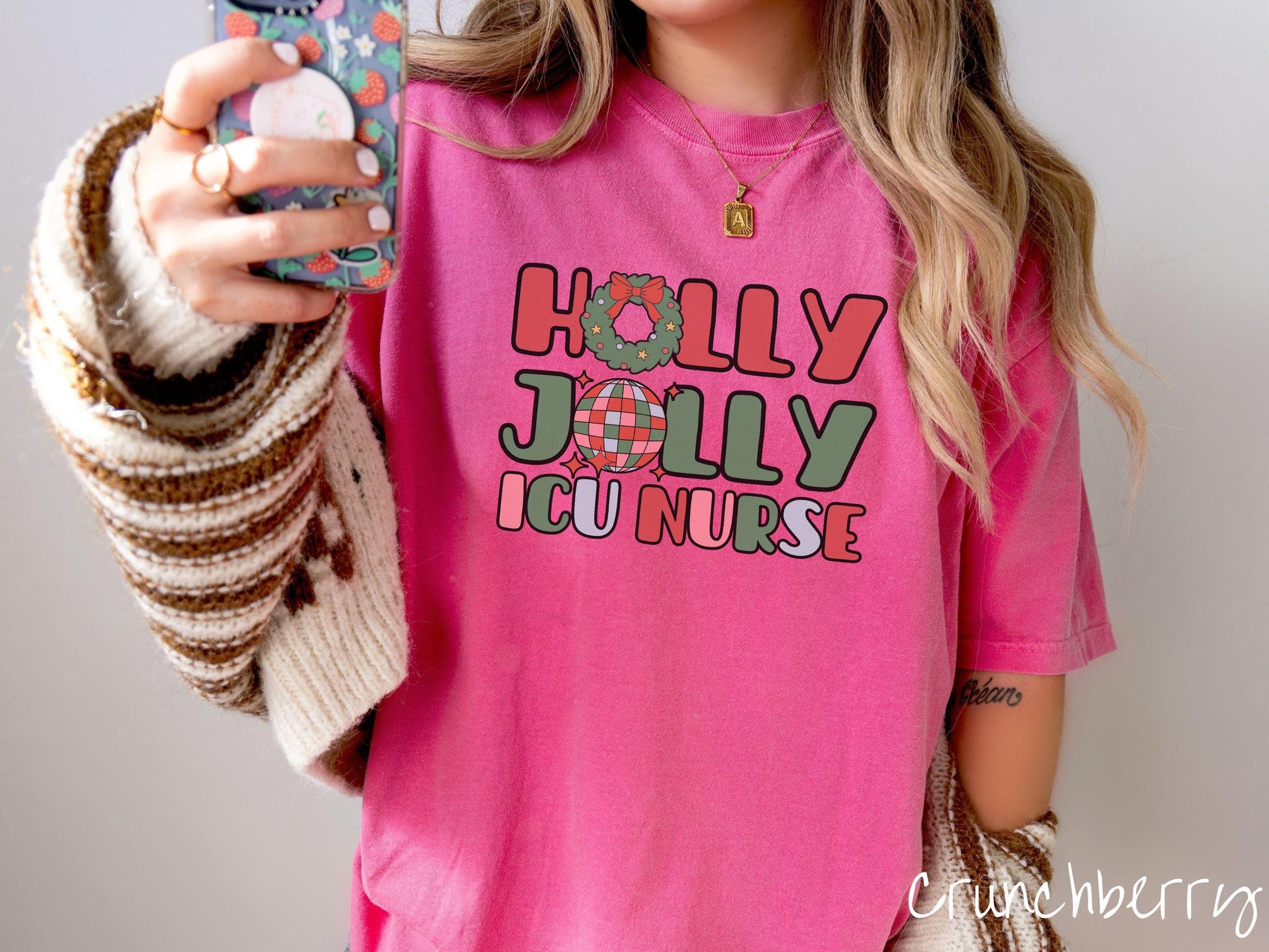 A woman wearing a cute, vintage crunchberry colored Comfort Colors T-shirt with the text Holly Jolly ICU Nurse in red, green, pink, and blue font. The letter O in Holly is a Christmas wreath and the letter O in Jolly is a multi-colored disco ball.