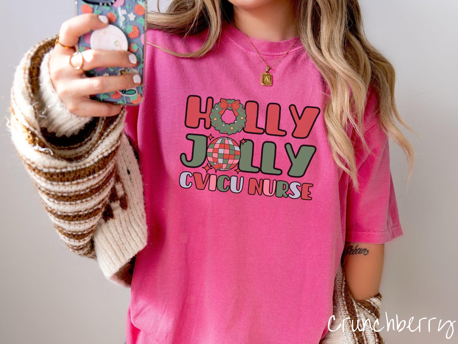 A woman wearing a cute, vintage crunchberry colored Comfort Colors T-shirt with the text Holly Jolly CVICU Nurse in red, green, pink, and blue font. The letter O in Holly is a Christmas wreath and the letter O in Jolly is a multi-colored disco ball.
