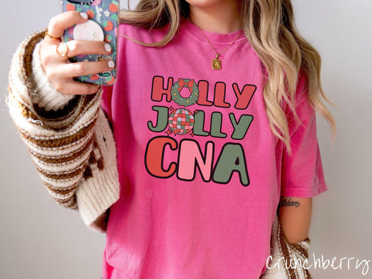 A woman wearing a cute, vintage crunchberry colored Comfort Colors T-shirt with the text Holly Jolly CNA in red, green, pink, and blue font. The letter O in Holly is a Christmas wreath and the letter O in Jolly is a multi-colored disco ball.