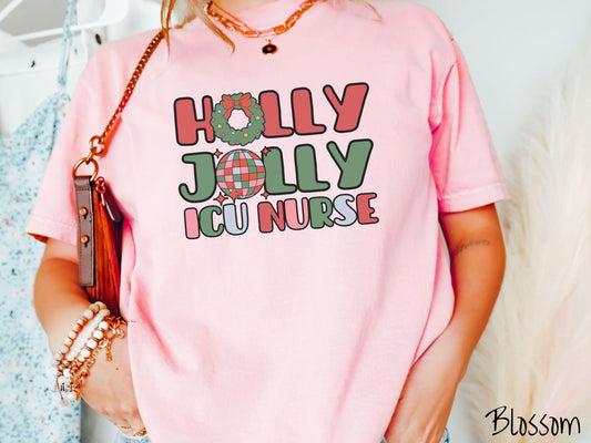 A woman wearing a cute, vintage blossom colored Comfort Colors T-shirt with the text Holly Jolly ICU Nurse in red, green, pink, and blue font. The letter O in Holly is a Christmas wreath and the letter O in Jolly is a multi-colored disco ball.