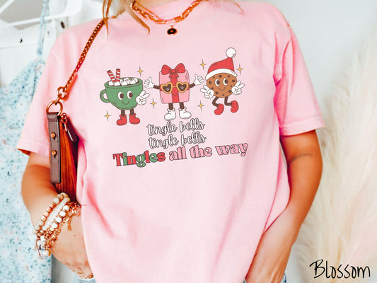 A cute, vintage blossom colored Comfort Colors T-shirt with the text tnigle bells, tingle bells, tingles all the way. Above are a green cup of hot cocoa holding a peace sign, a red present holding two thumbs up, and a cookie holding up peace signs.
