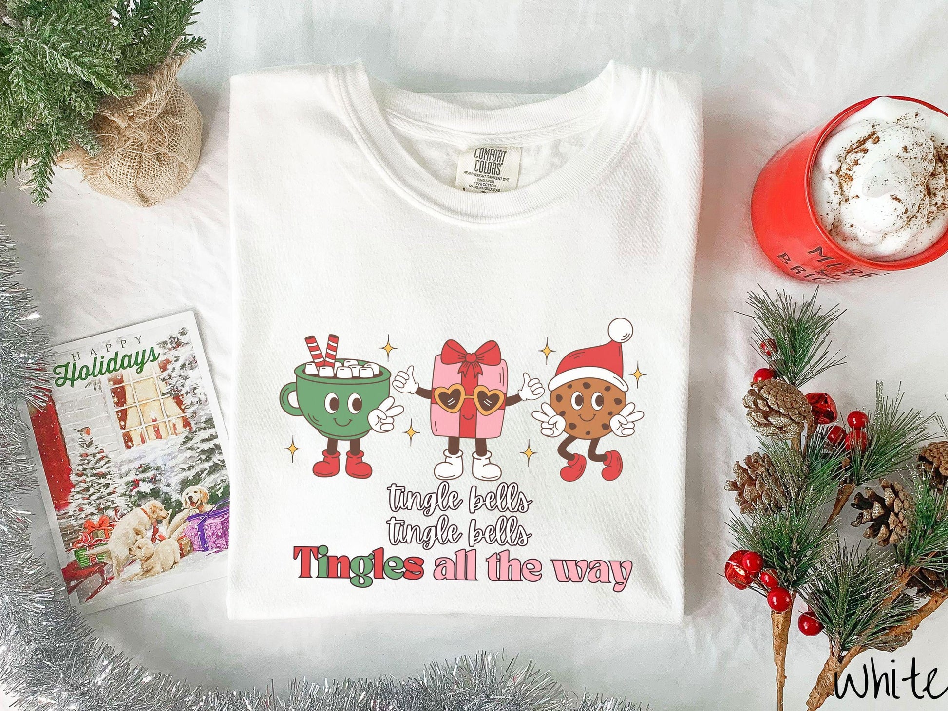 A cute, vintage white colored Comfort Colors T-shirt with the text tnigle bells, tingle bells, tingles all the way. Above are a green cup of hot cocoa holding a peace sign, a red present holding two thumbs up, and a cookie holding up peace signs.