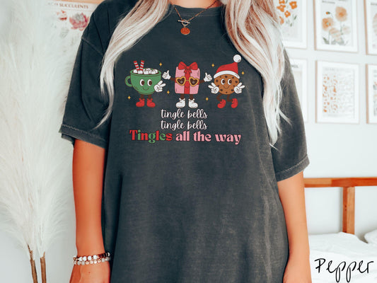 A cute, vintage pepper colored Comfort Colors T-shirt with the text tnigle bells, tingle bells, tingles all the way. Above are a green cup of hot cocoa holding a peace sign, a red present holding two thumbs up, and a cookie holding up peace signs.