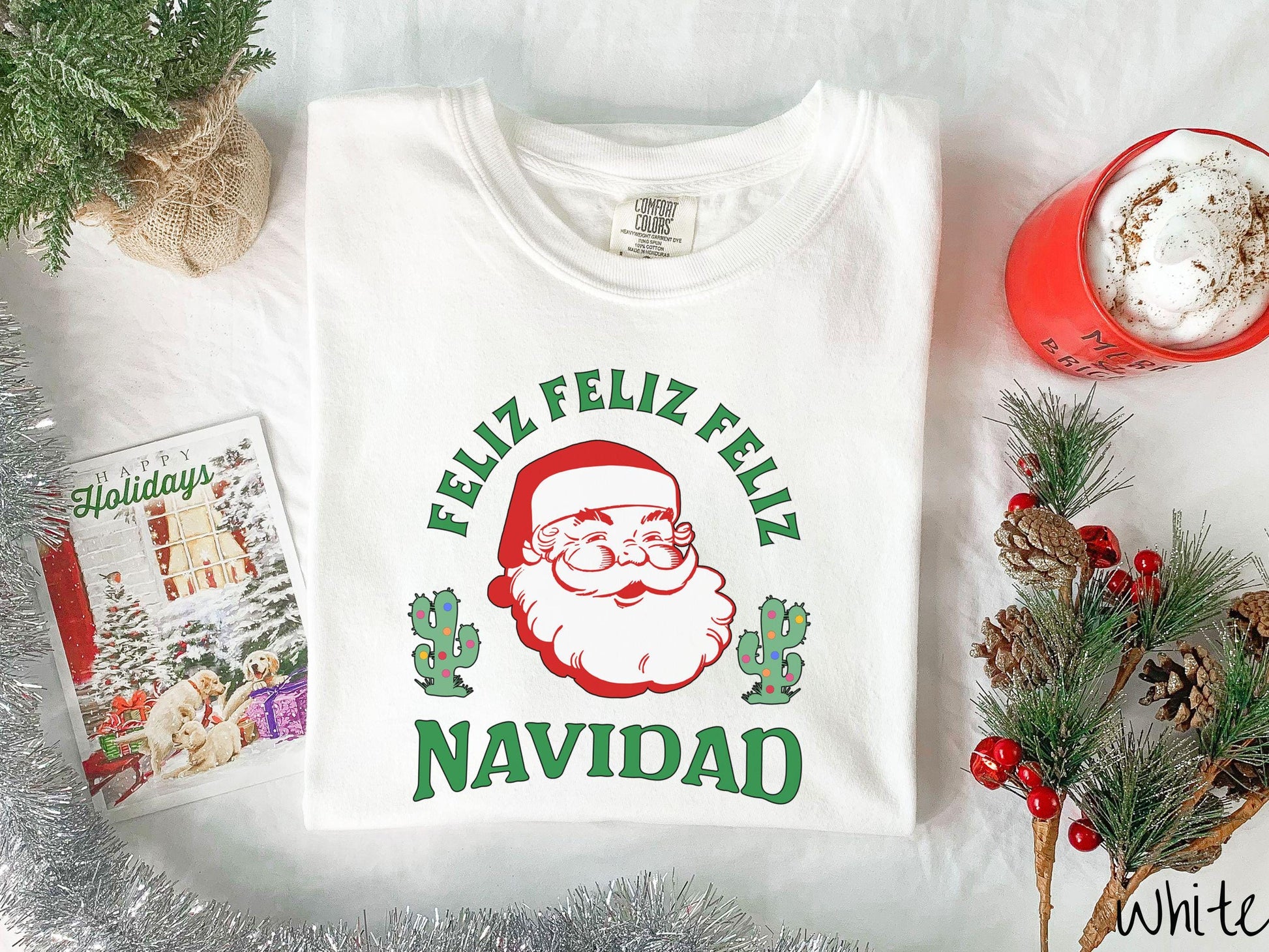 This festive white colored Comfort Colors t-shirt featuring a smiling santa clause between two green cacti covered in colorful Christmas lights and the phrase, 'Feliz feliz feliz Navidad' is perfect for spreading holiday cheer.